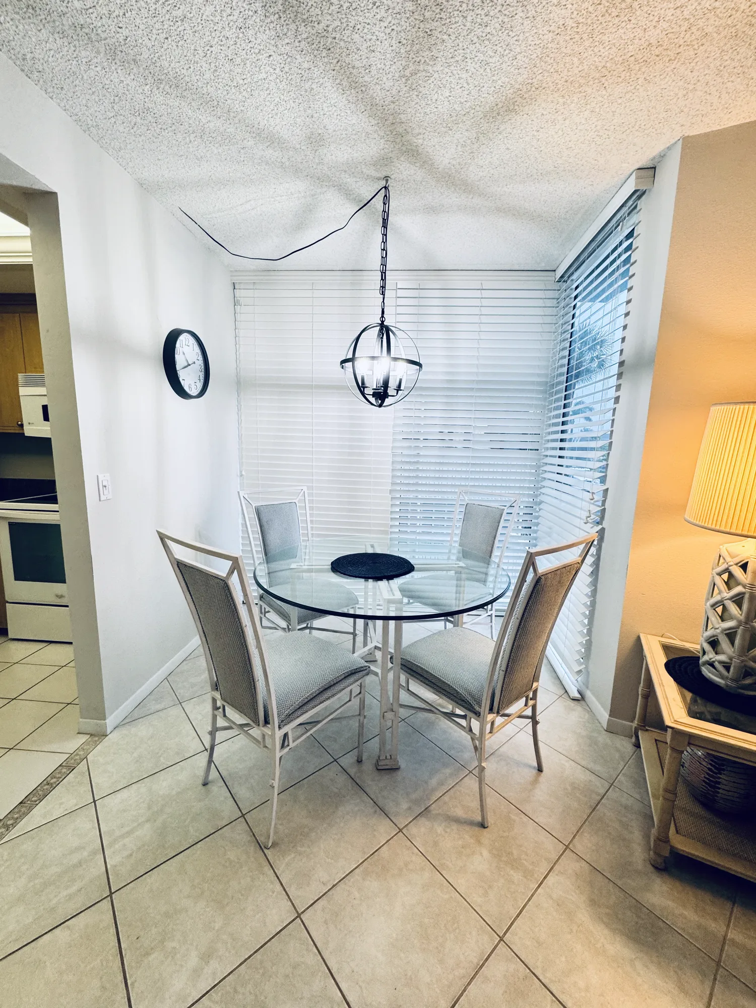Dining nook with a round glass table, four chairs and a pendant light, beside a window with blinds.