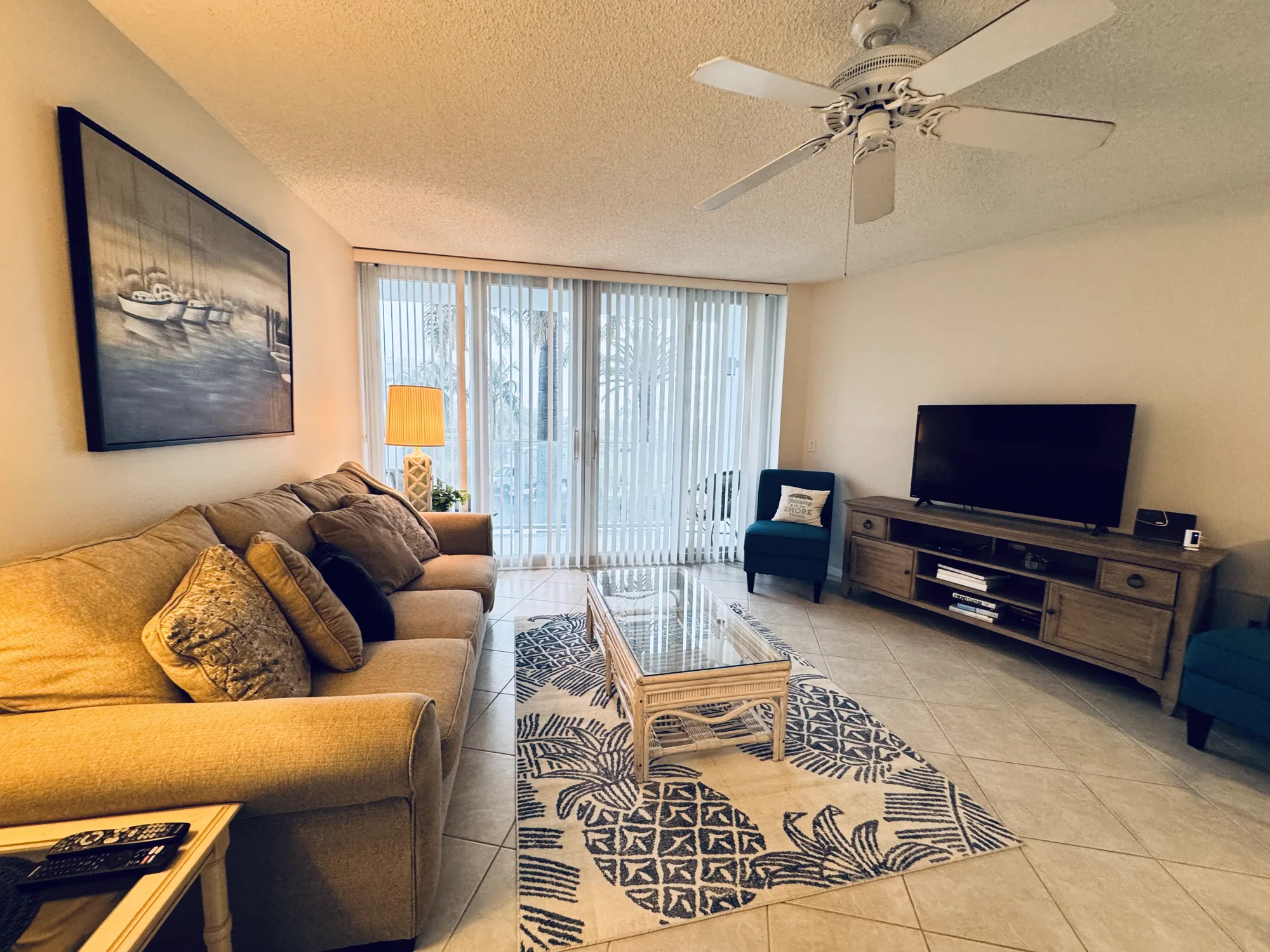 Living room with a tan sofa, a glass coffee table on a patterned rug, a television on a wood console, and sliding doors to the balcony with palms outside.
