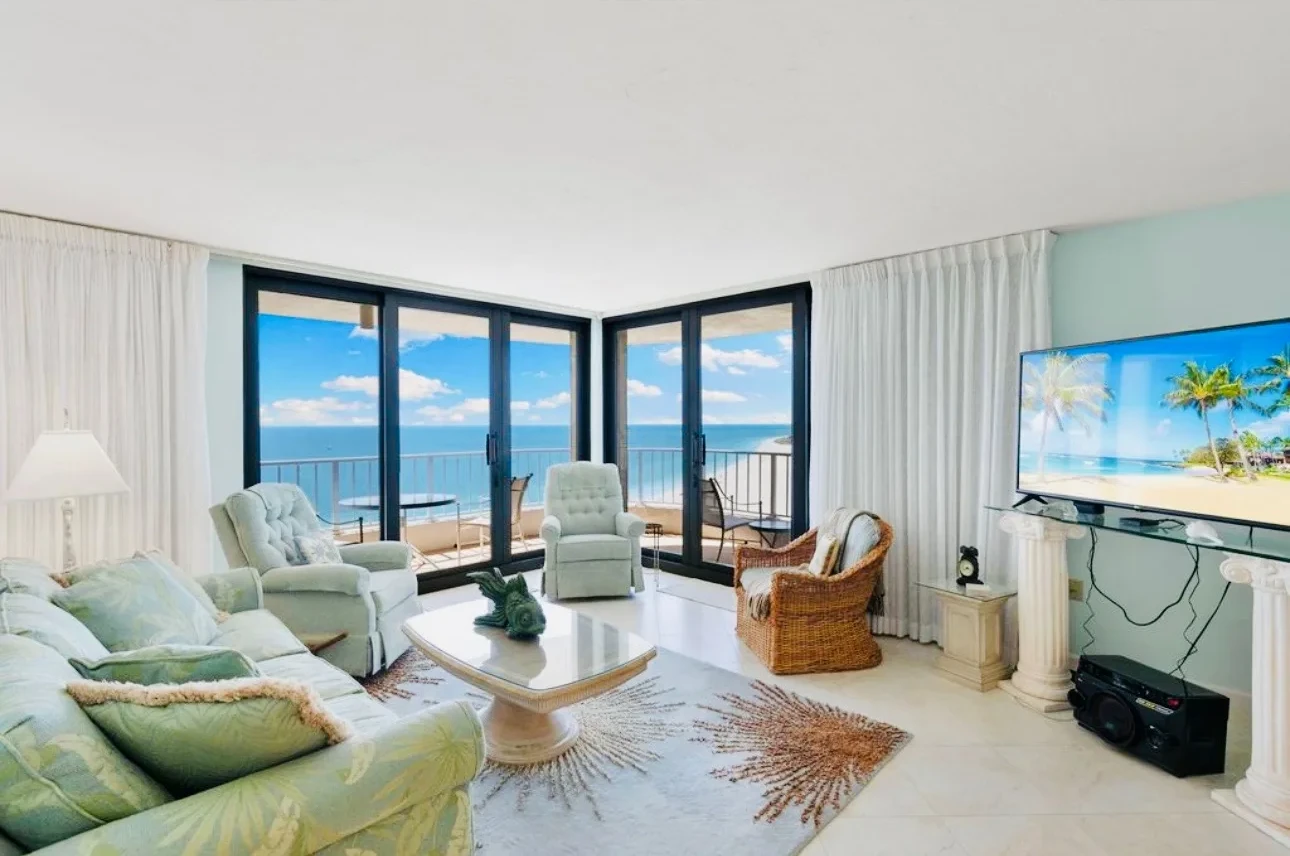 Living room seen from the sofa: pale green sofa and armchairs, a glass coffee table, a television on the right, and the beach through the sliding doors.