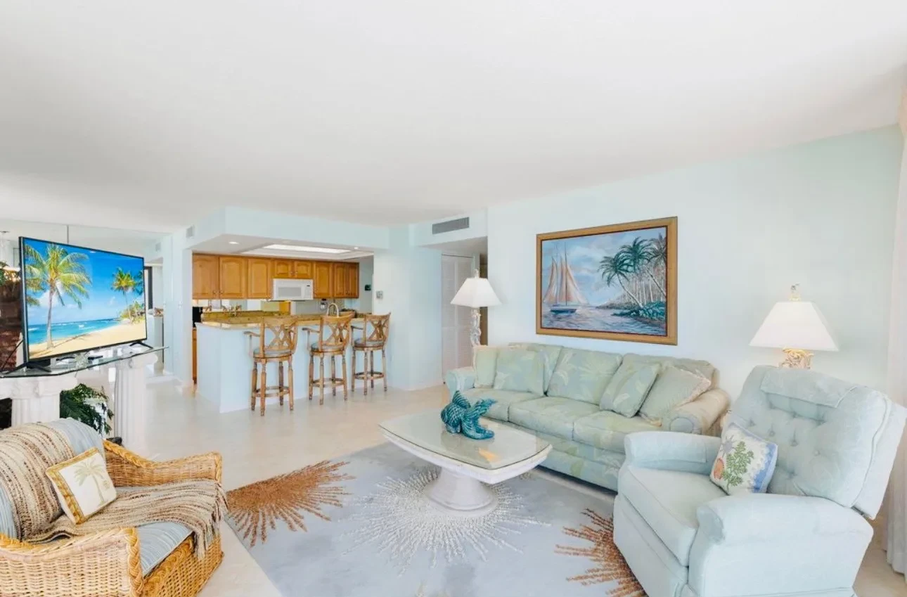 Wide view of the living room with the kitchen and breakfast bar behind it, pale green furniture and tile floors.