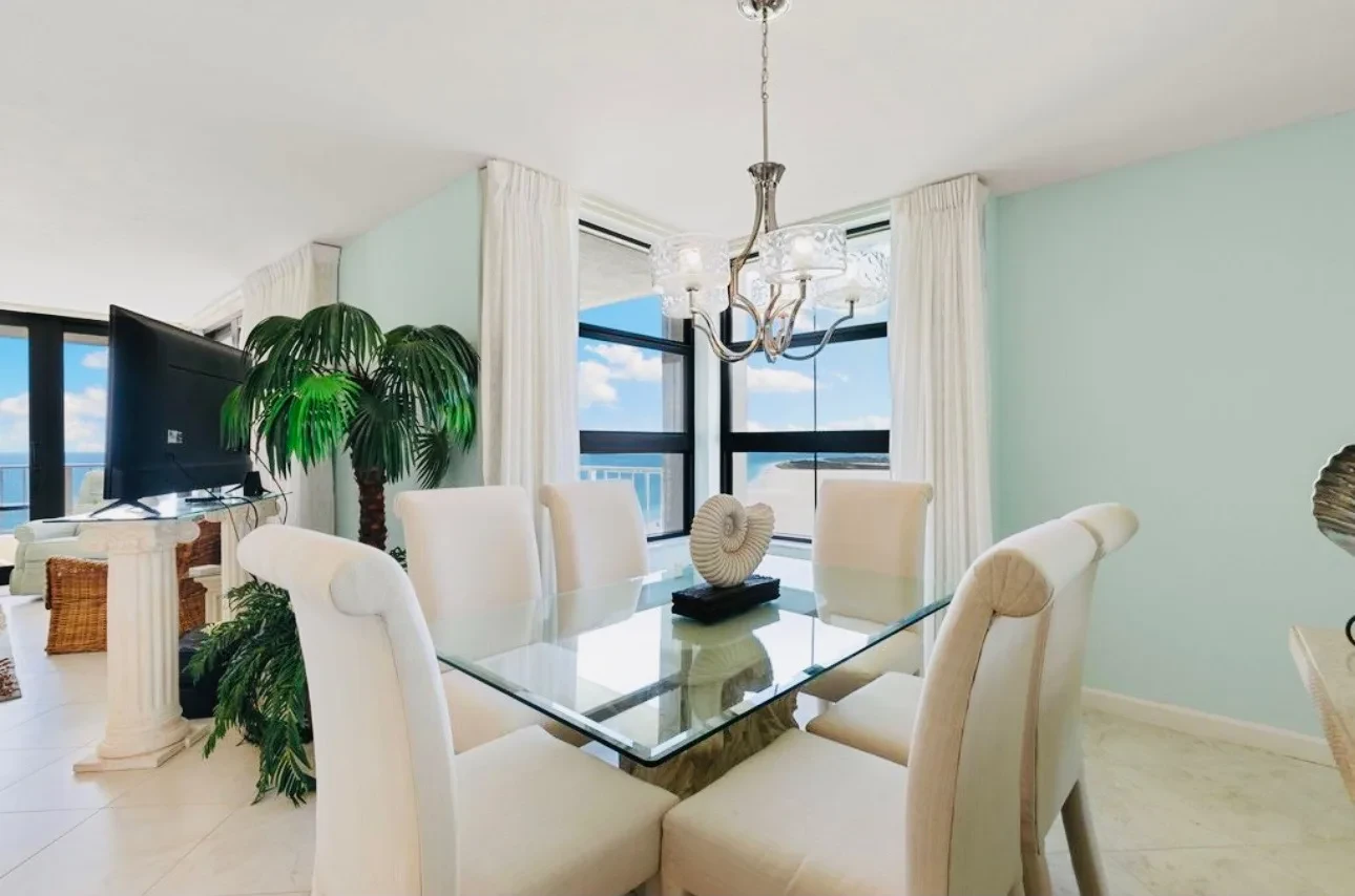 Dining area with a glass table and six upholstered chairs, a chandelier above, and windows with the beach behind.
