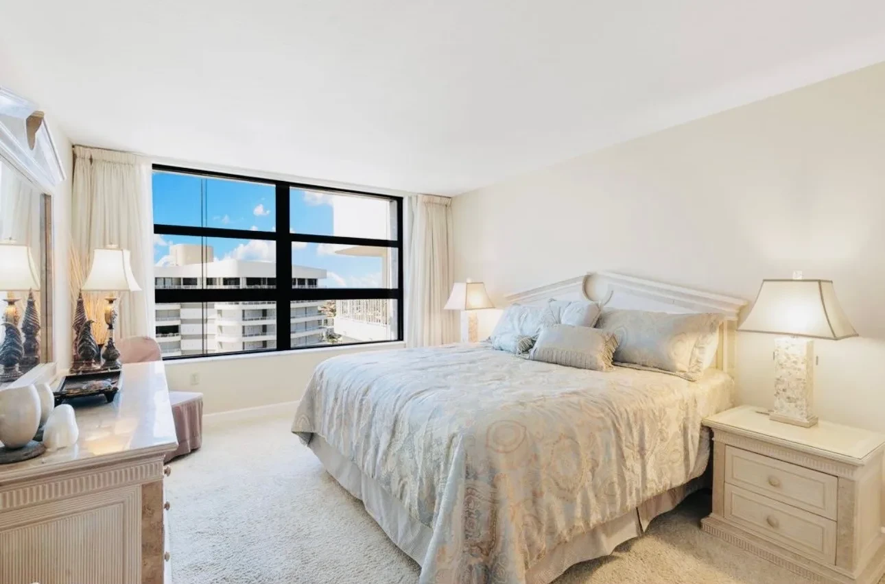 Bedroom with a large bed in cream linens, two bedside lamps, and a wide window looking out over neighbouring buildings toward the beach.