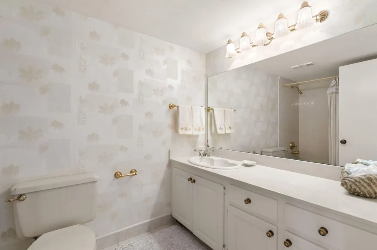 Bathroom with a long white vanity, a wide mirror and light patterned wallpaper.