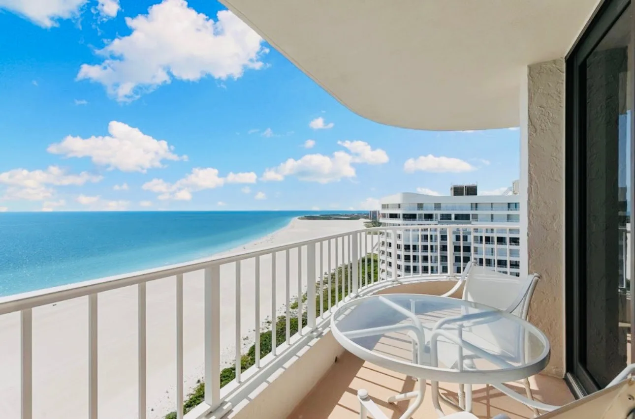 View from the wraparound deck, looking along the white beach with the Gulf on the left and neighbouring buildings on the right. A glass table and two chairs sit on the deck.